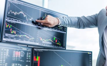 Businessmen trading stocks online. Stock brokers looking at graphs, indexes and numbers on multiple computer screens. Colleagues in discussion in traders office. Business success concept.